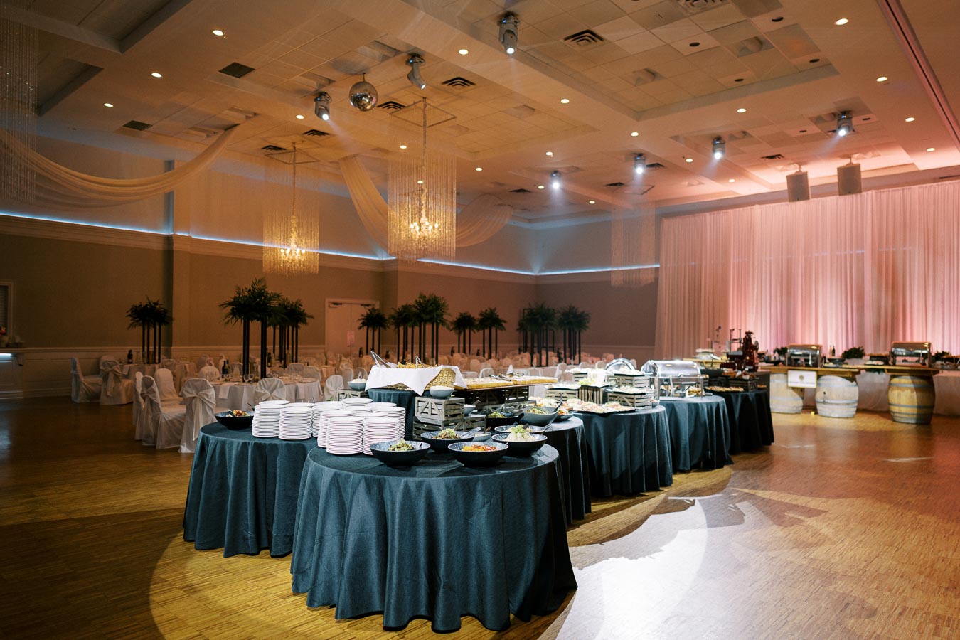 Elegant banquet hall with buffet setup featuring round tables covered with black tablecloths, plates, and a variety of dishes, surrounded by decorated chairs and ambient lighting perfect for weddings or special events.