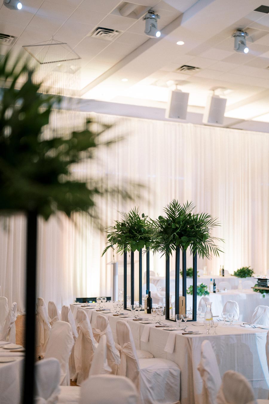 Elegant wedding reception with white-draped tables and chairs, adorned with tall greenery centerpieces, in a softly lit venue setting.