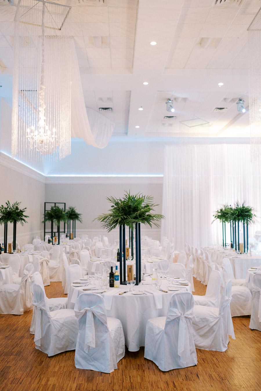 Luxurious wedding reception hall with elegant white draped tables and chairs, tall centerpieces with lush green plants, and a sparkling chandelier hanging from the ceiling.