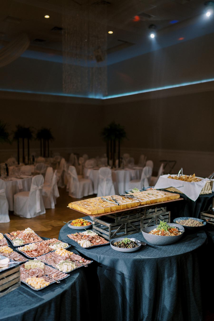 Elegant banquet setup in a dimly lit reception hall, featuring a variety of gourmet appetizers on black tablecloths, including charcuterie, cheese platters, and assorted breads, ready for guests.