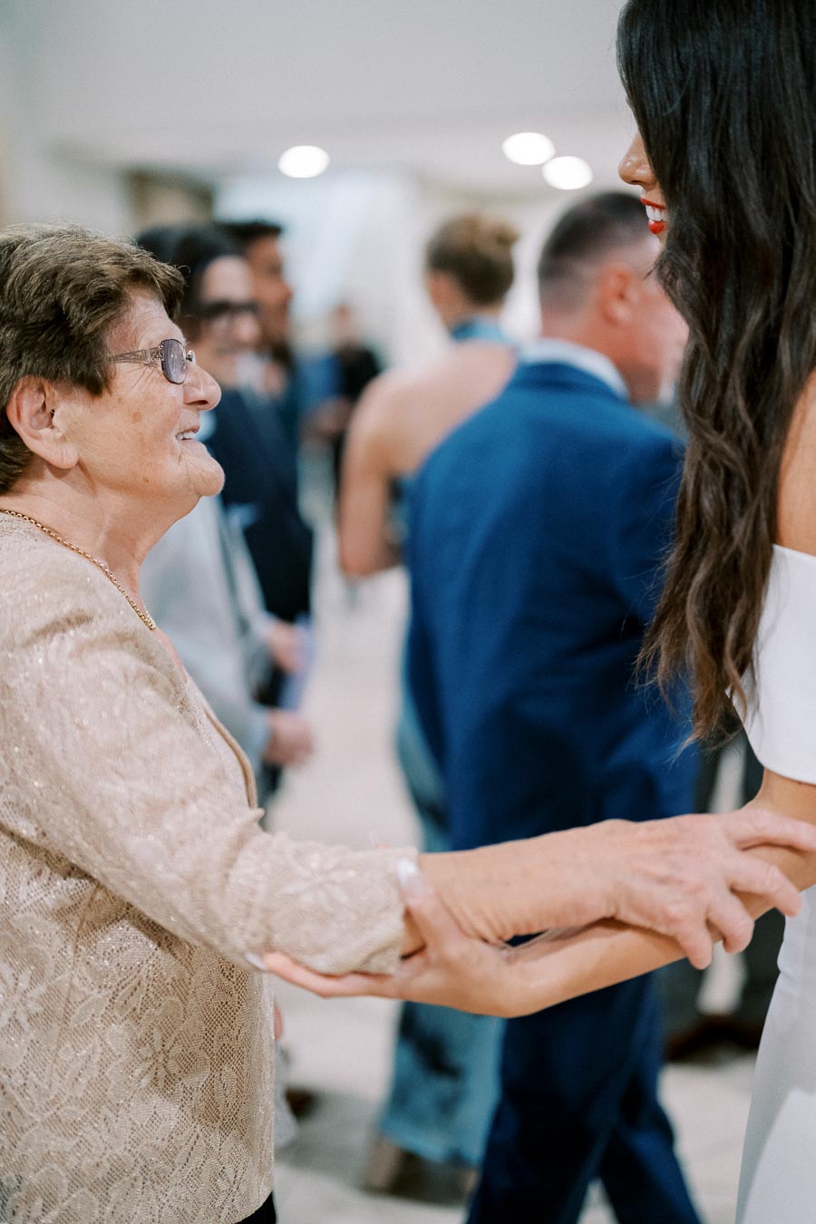 Elderly woman in a beige dress holding hands with a younger woman in a white dress at a social gathering.