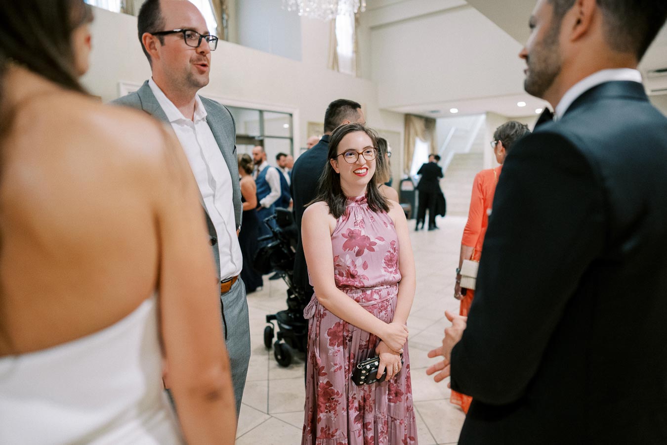 A group of people at a formal event, engaged in conversation. A woman in a floral dress is smiling, while others in suits and evening attire are talking in a well-lit, elegant venue.
