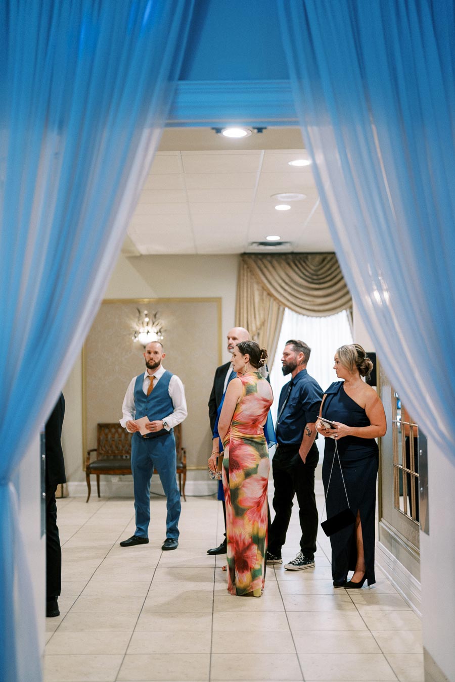 Elegant wedding guests in formal attire gathered in a beautifully decorated venue with blue drapes, preparing for a special event.