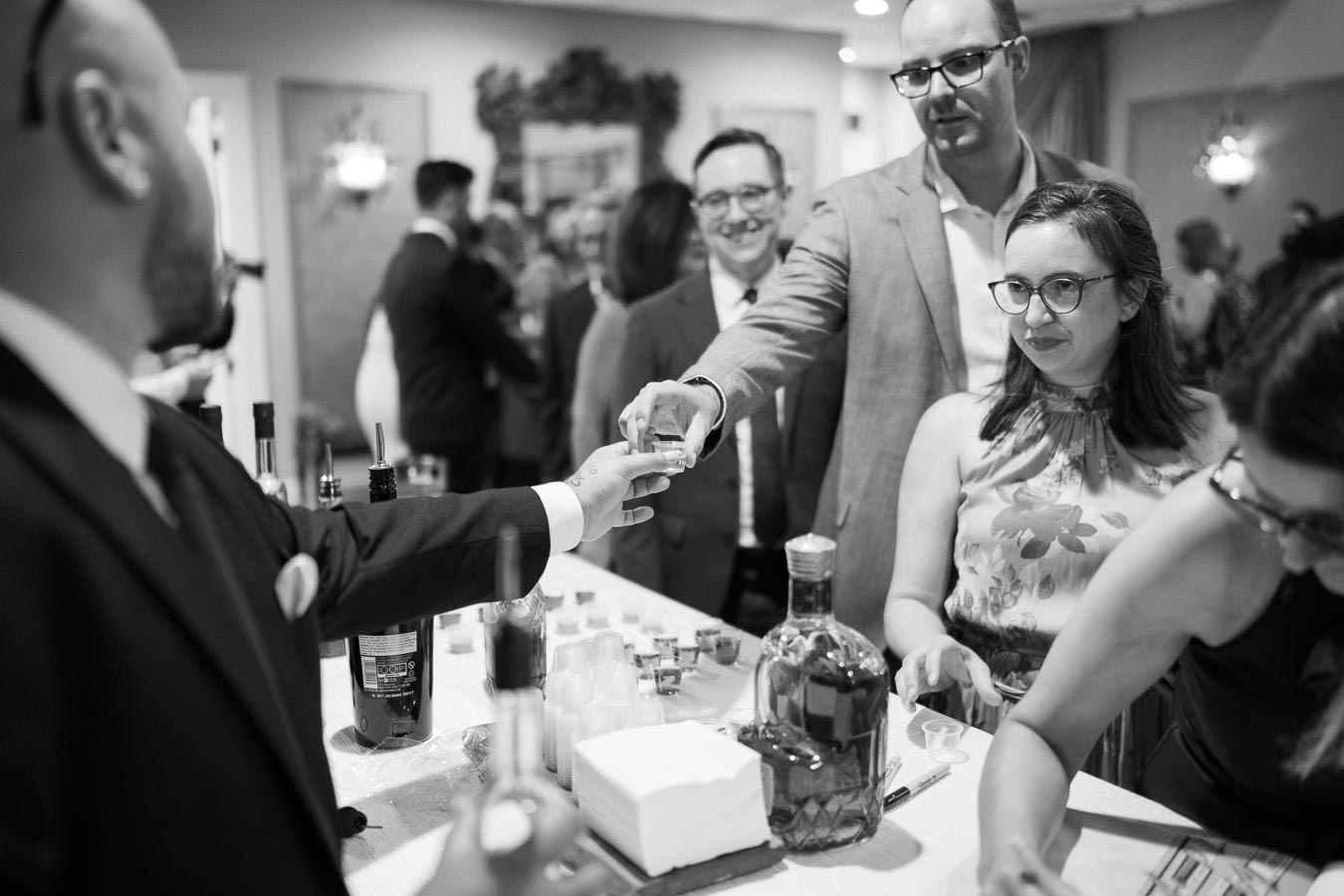 A black and white image of people gathered around a bar, with a man serving drinks and others socializing, creating a lively atmosphere at an event reception. Bottles and glasses are visible on the counter.
