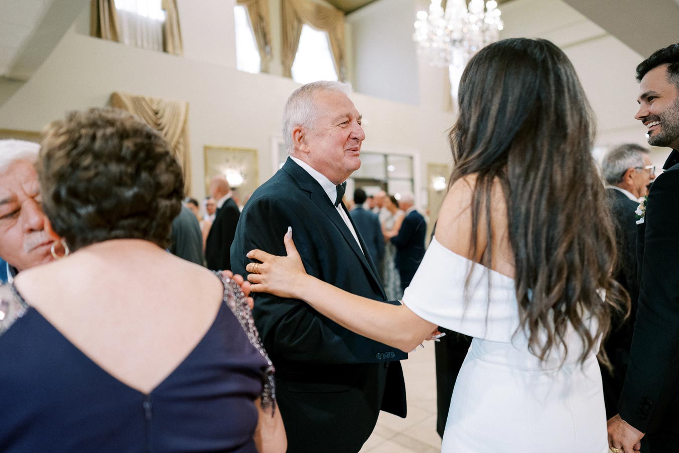 A group of people socializing at a formal event, with a woman in a white dress interacting with an older man in a suit, surrounded by elegantly dressed guests in a well-lit room.