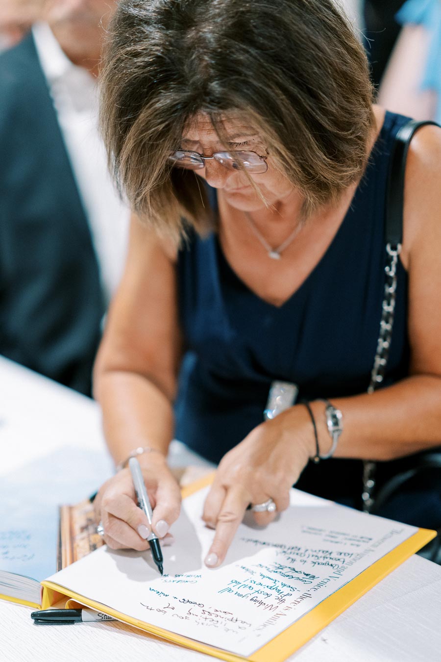 A woman with shoulder-length hair and glasses writing notes in a book with a pen, focused on her writing, at a public signing event.