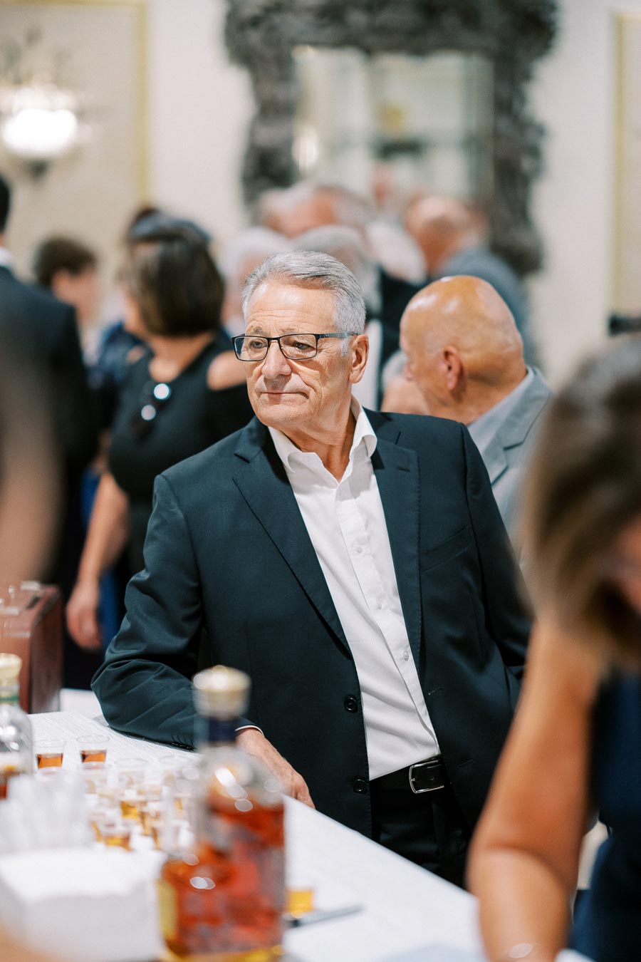 Elegant man in a suit at a social gathering with drinks on the table, surrounded by a crowd in a sophisticated indoor setting.