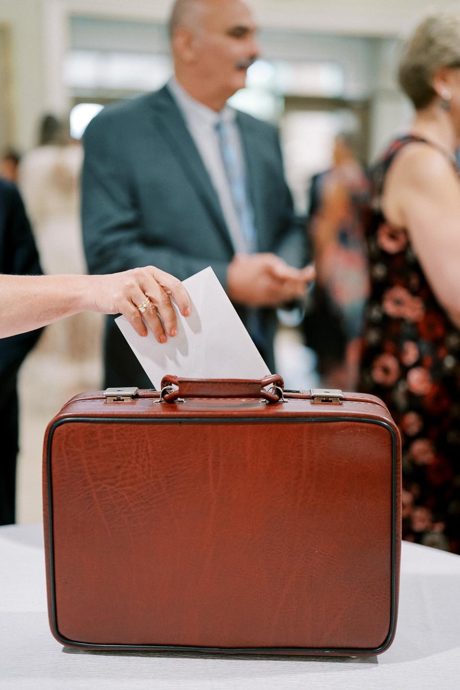 Person placing an envelope into a vintage leather suitcase, possibly symbolizing travel or an event collection, with people in formal attire in the background.
