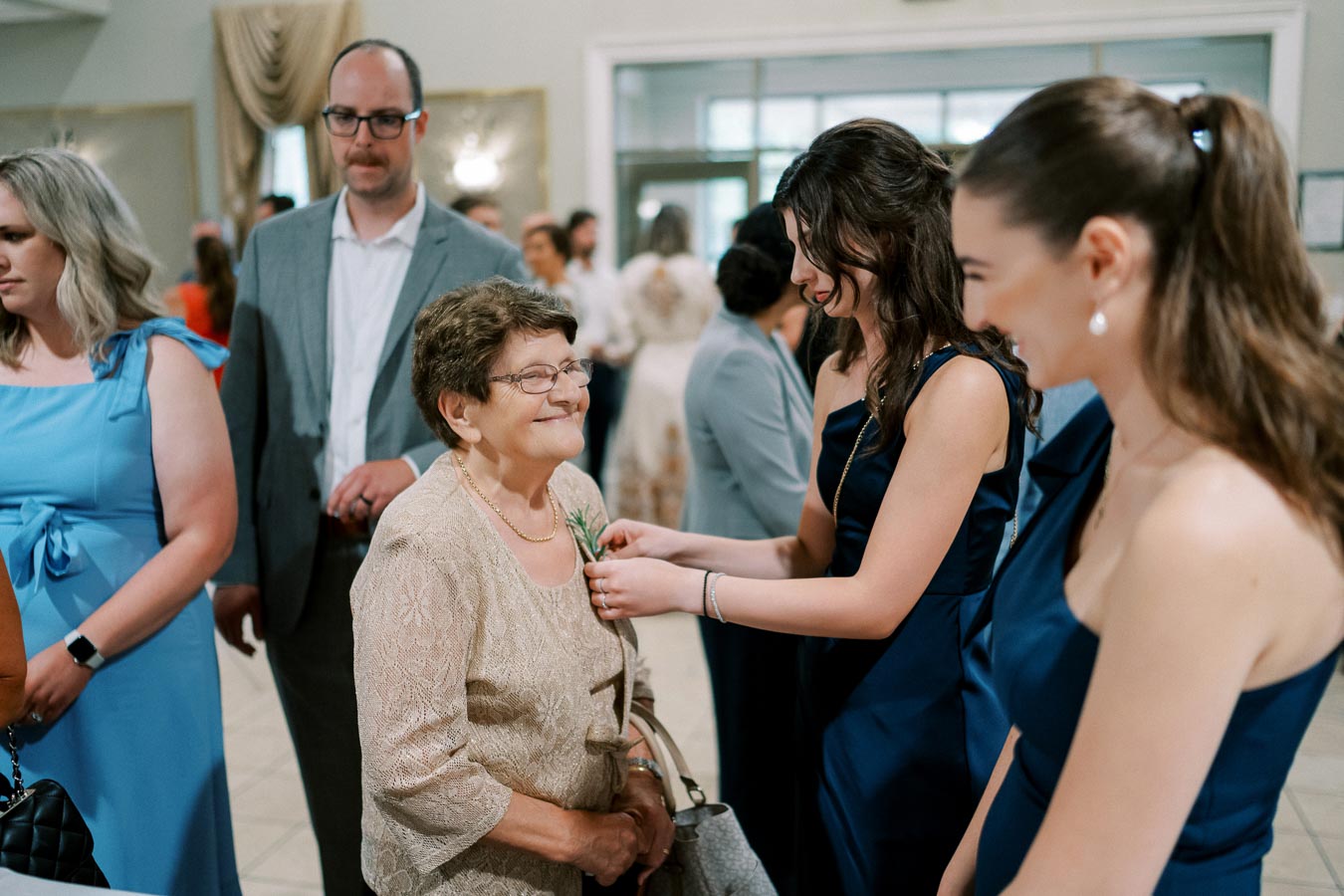 A joyful elderly woman wearing a lace dress being helped with a boutonniere by a young woman in a blue dress at a social event, surrounded by guests in formal attire.