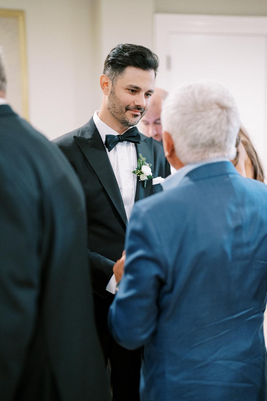 A man in a black tuxedo, wearing a white shirt and bow tie, stands inside a room talking to another man in a blue suit. The setting appears formal, possibly at a wedding or similar event