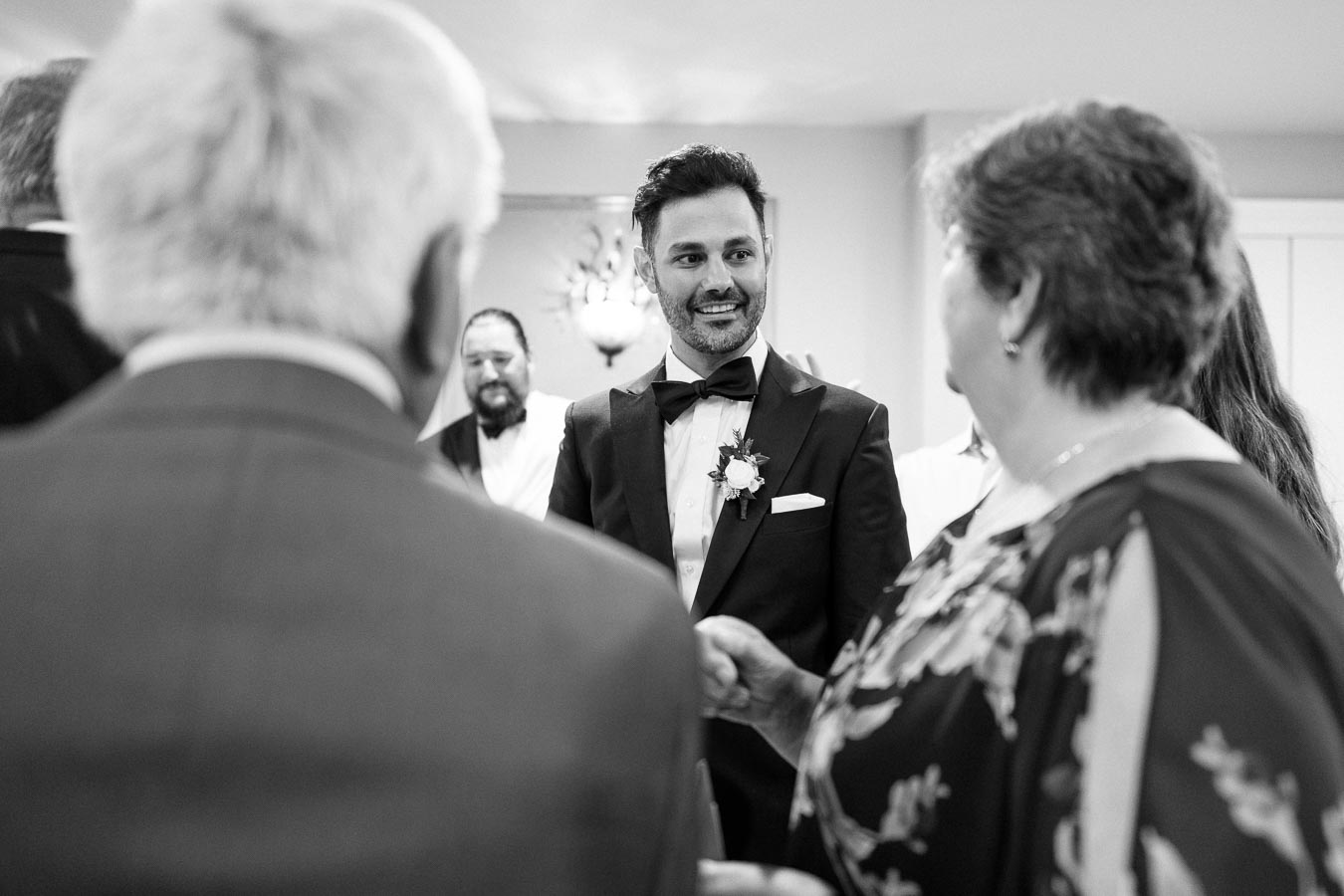 Black and white photo of a smiling groom in a tuxedo interacting with guests at a wedding reception, highlighting a joyful and elegant atmosphere.