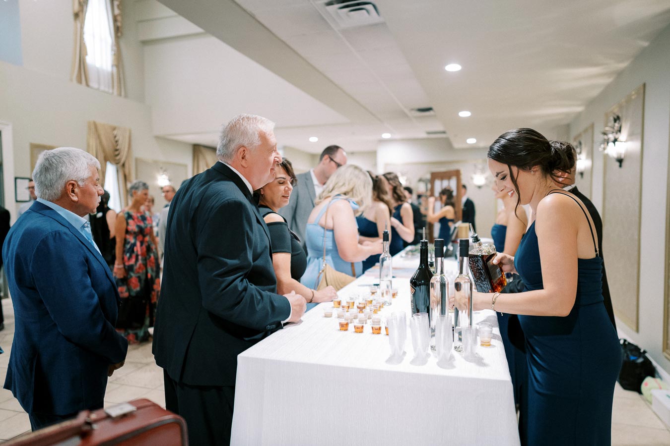 Guests in formal attire gather around a bar table at a wedding reception, with bottles of wine and glasses arranged neatly, in a bright and elegantly decorated venue.