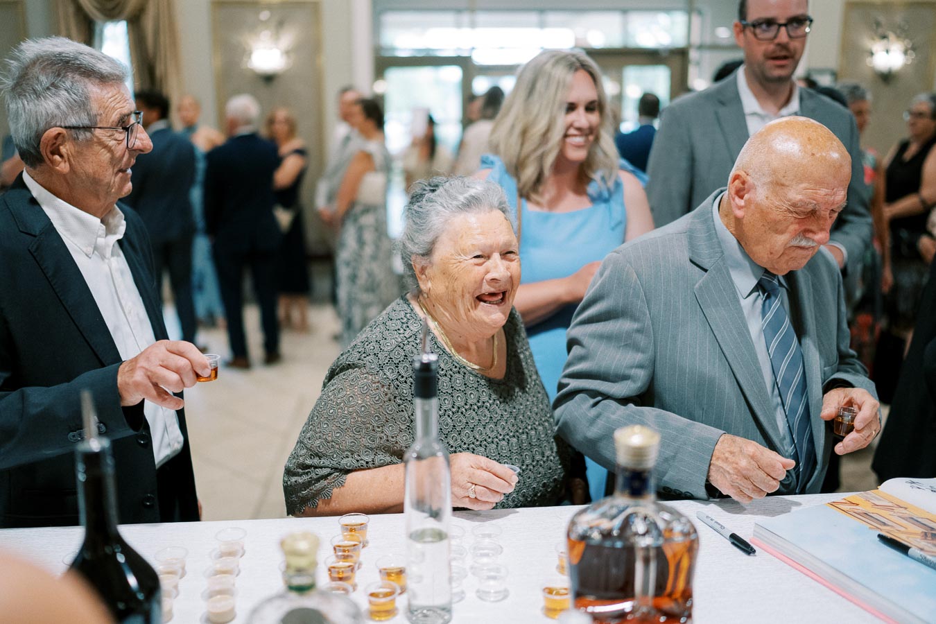 Elderly couple enjoying a social gathering at a reception, engaging in conversation with friends while holding small glasses of liquor, with bottles displayed on a table in the foreground.