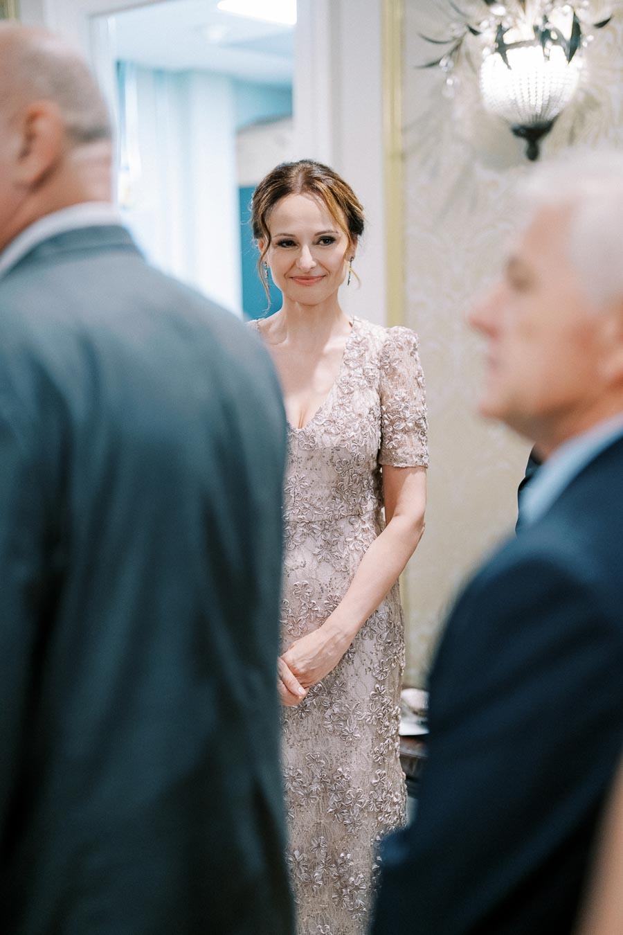 Elegant woman in a lace dress smiling at a formal gathering, with blurred figures in the foreground at a sophisticated event.