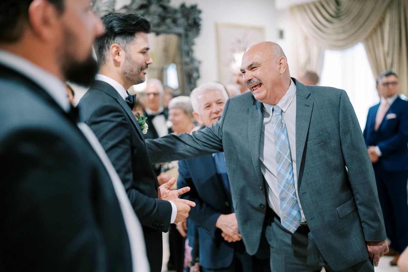 A joyful moment captured at a formal event, featuring a group of well-dressed individuals in suits, with two men in conversation and laughter, set against a sophisticated indoor backdrop.