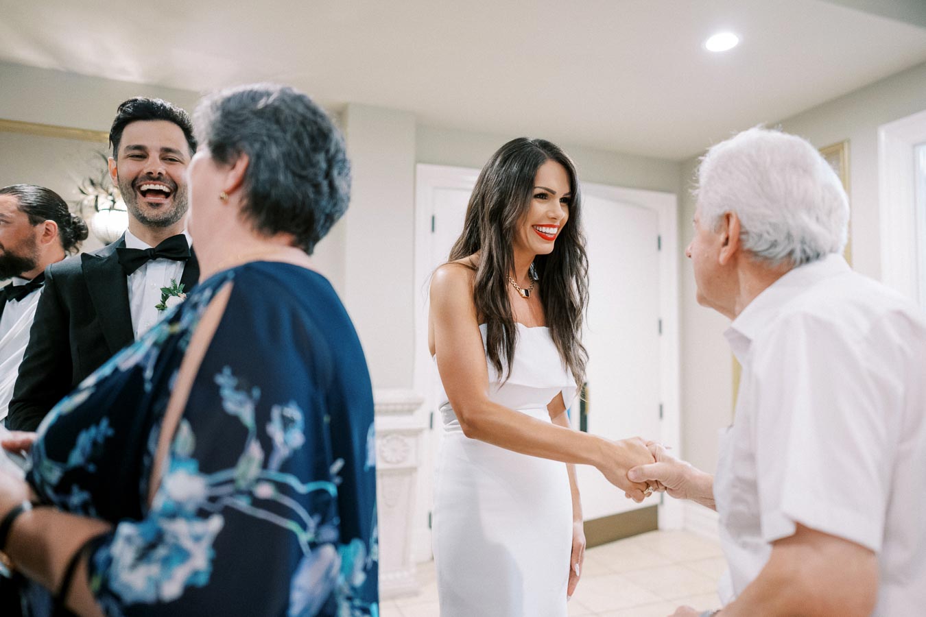 Guests at a wedding reception, with a woman in a white dress smiling and shaking hands with an elderly man, while others chat in the background.
