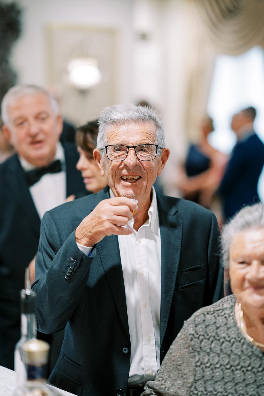 Elderly man in a suit enjoying a drink at a social gathering, surrounded by friends and family in a warmly lit room.