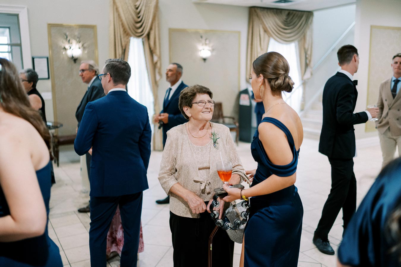 Guests mingling at a formal event, with an elderly woman in a beige outfit conversing with a young woman in a navy blue dress. Elegant interior decor with draped curtains and chandeliers in the background.