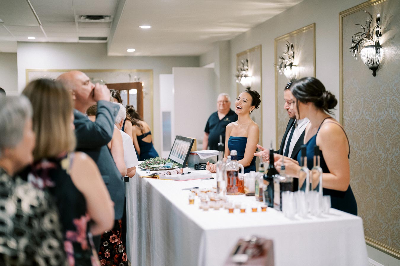 Guests enjoy drinks at a lively event bar, with bartenders serving beverages and attendees engaging happily in conversation in an elegant, well-lit venue.