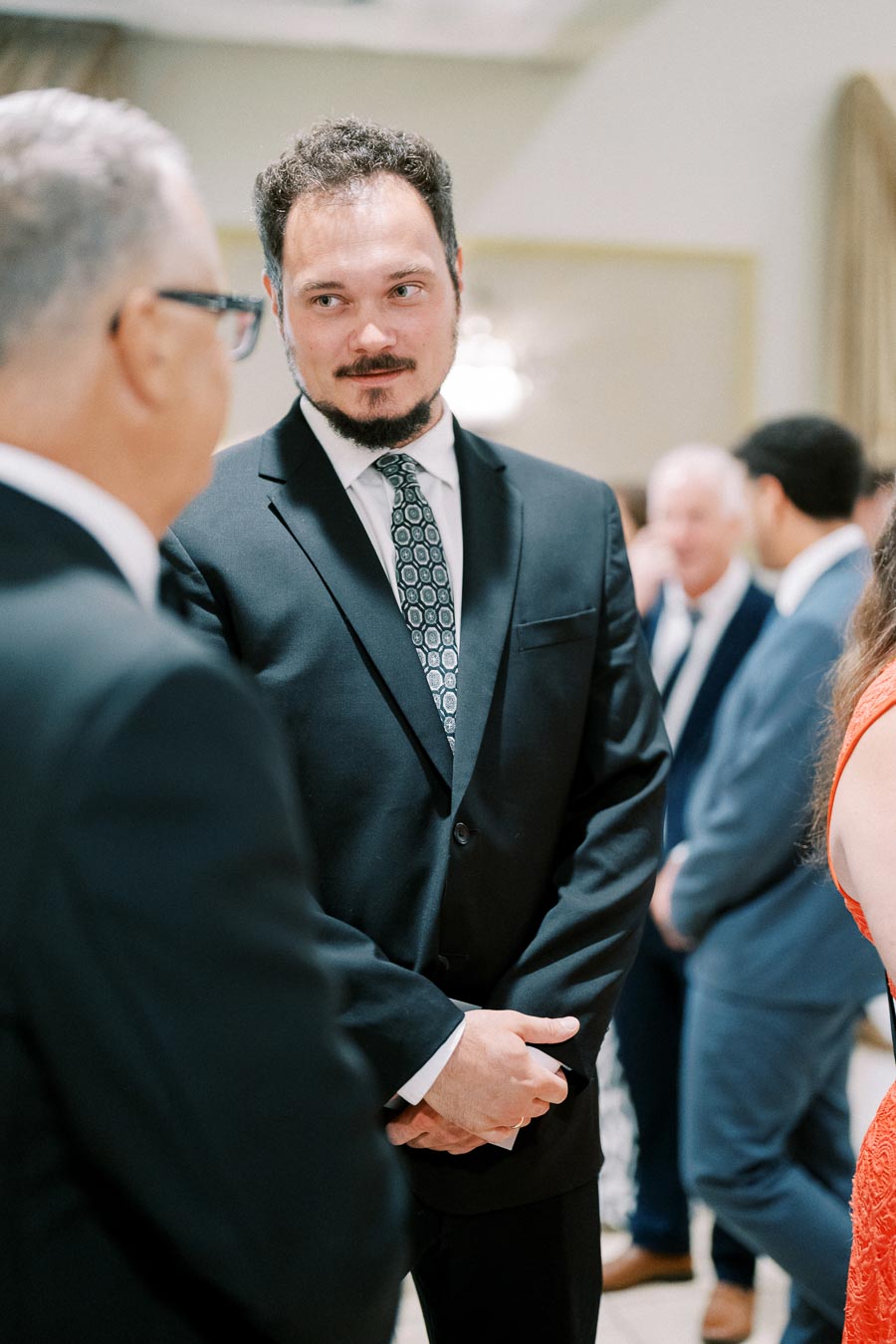 A man in a black suit and patterned tie engaged in conversation at a formal gathering, with people mingling in the background.