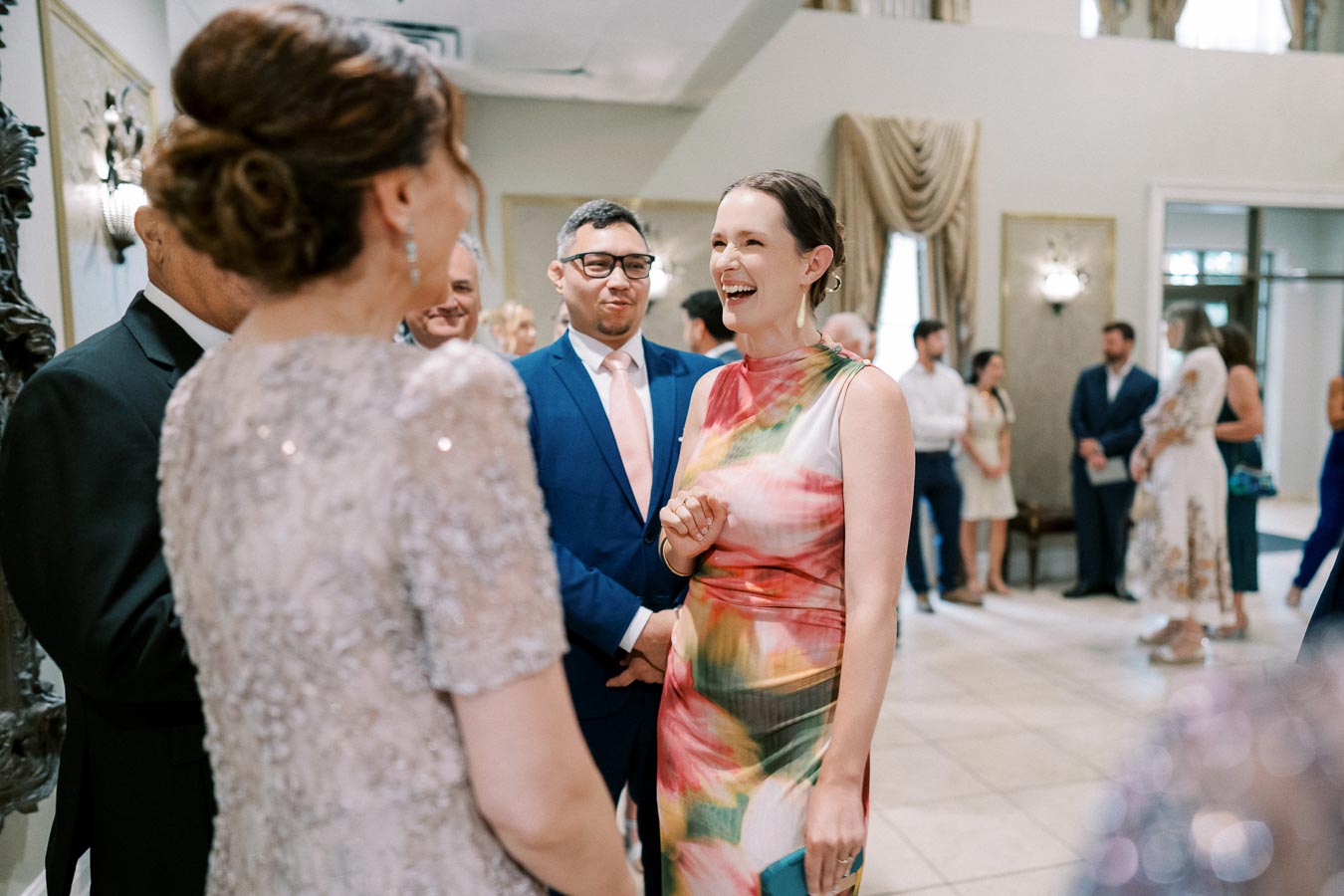 Group of elegantly dressed people socializing at a formal event, with a woman in a colorful dress smiling and interacting in a beautifully decorated reception area.