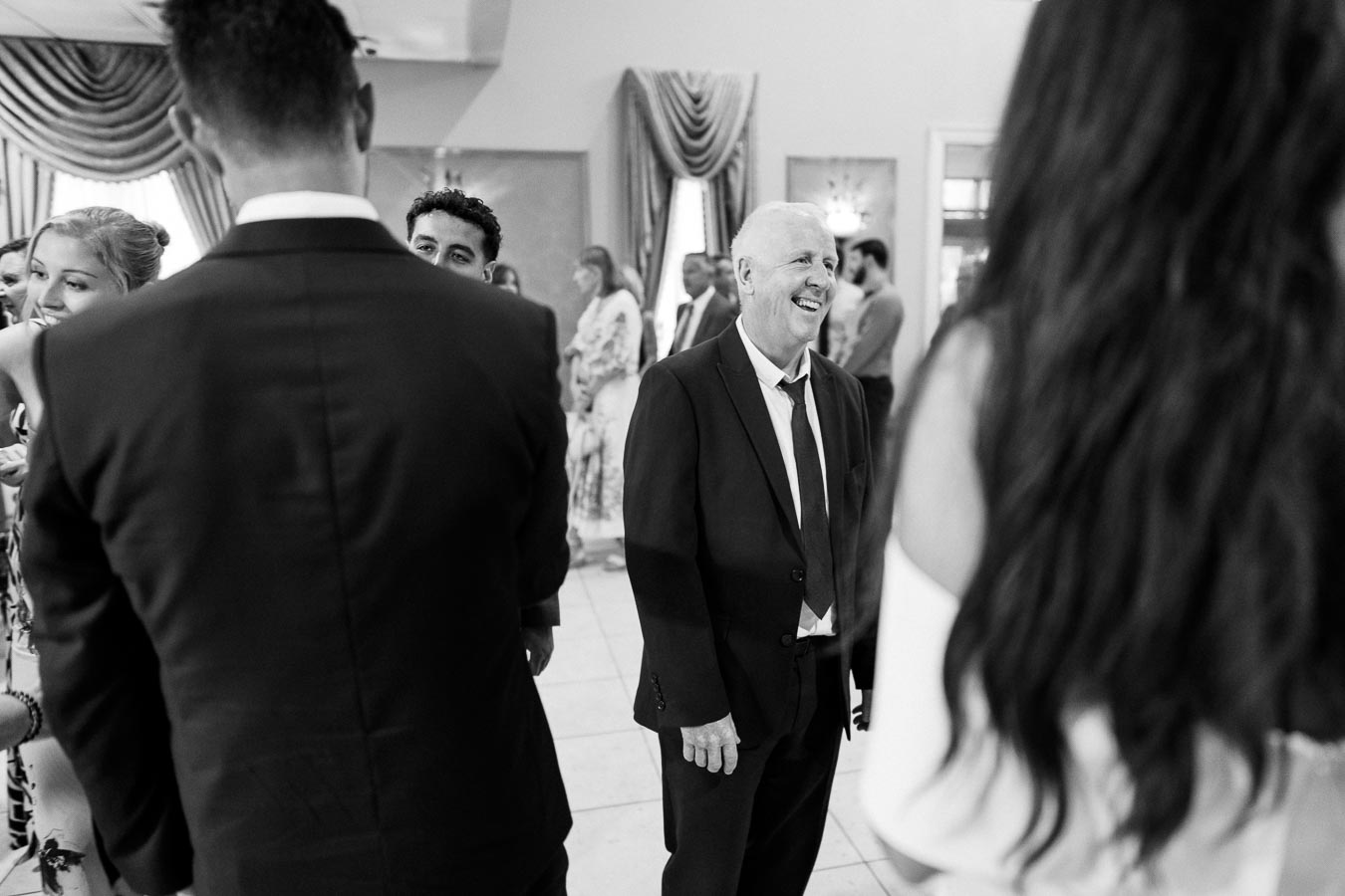 Black and white photo of people socializing at an indoor event, featuring a smiling man in a suit and tie surrounded by other guests. The background shows elegant drapery and a sophisticated atmosphere.