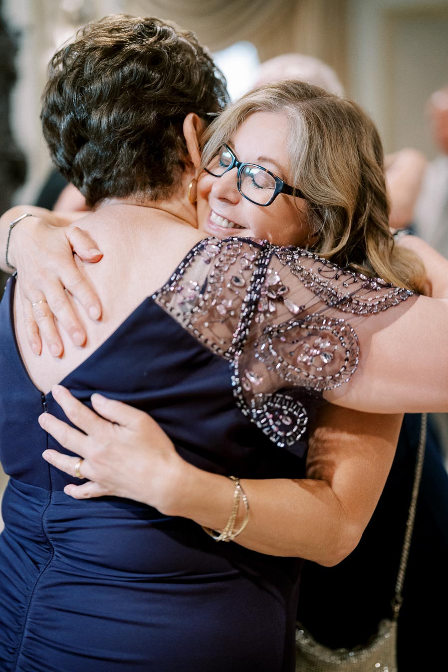 Elderly woman with glasses joyfully hugging another woman at a social event, both wearing elegant evening dresses.