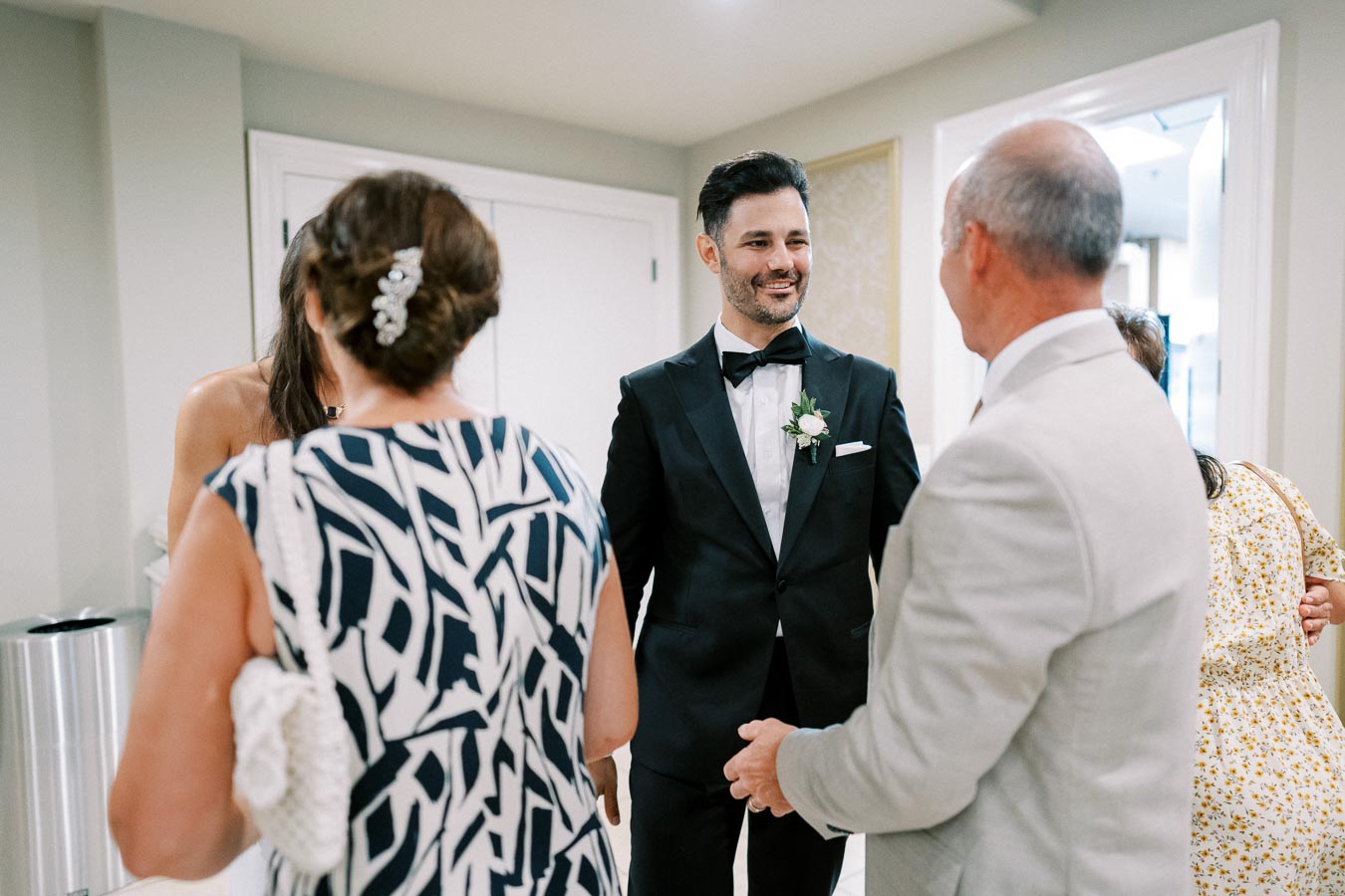 Groom in a black tuxedo smiling and greeting guests at a wedding reception.