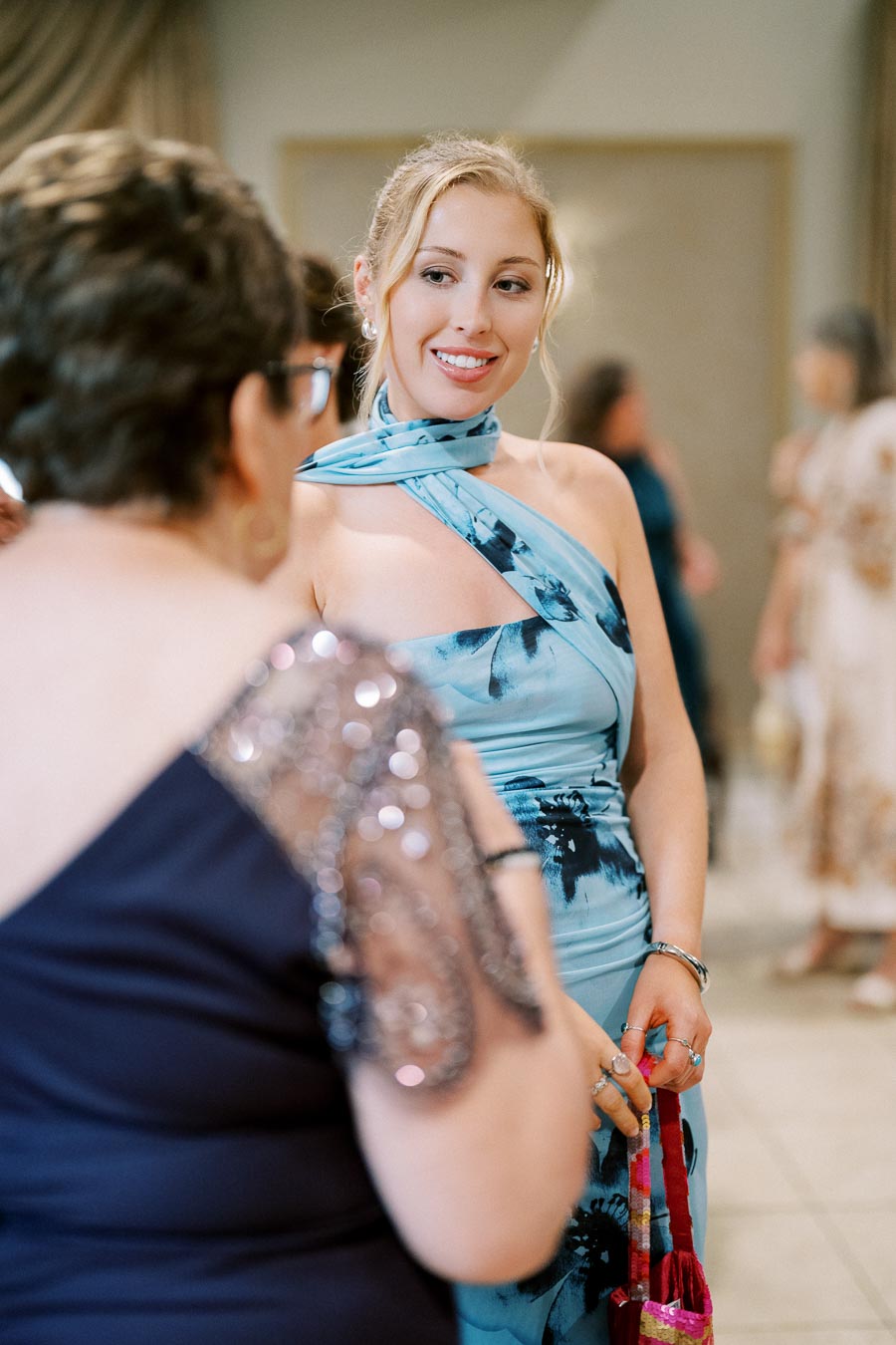 A woman in a blue floral dress smiles warmly while conversing with another person at a formal event.