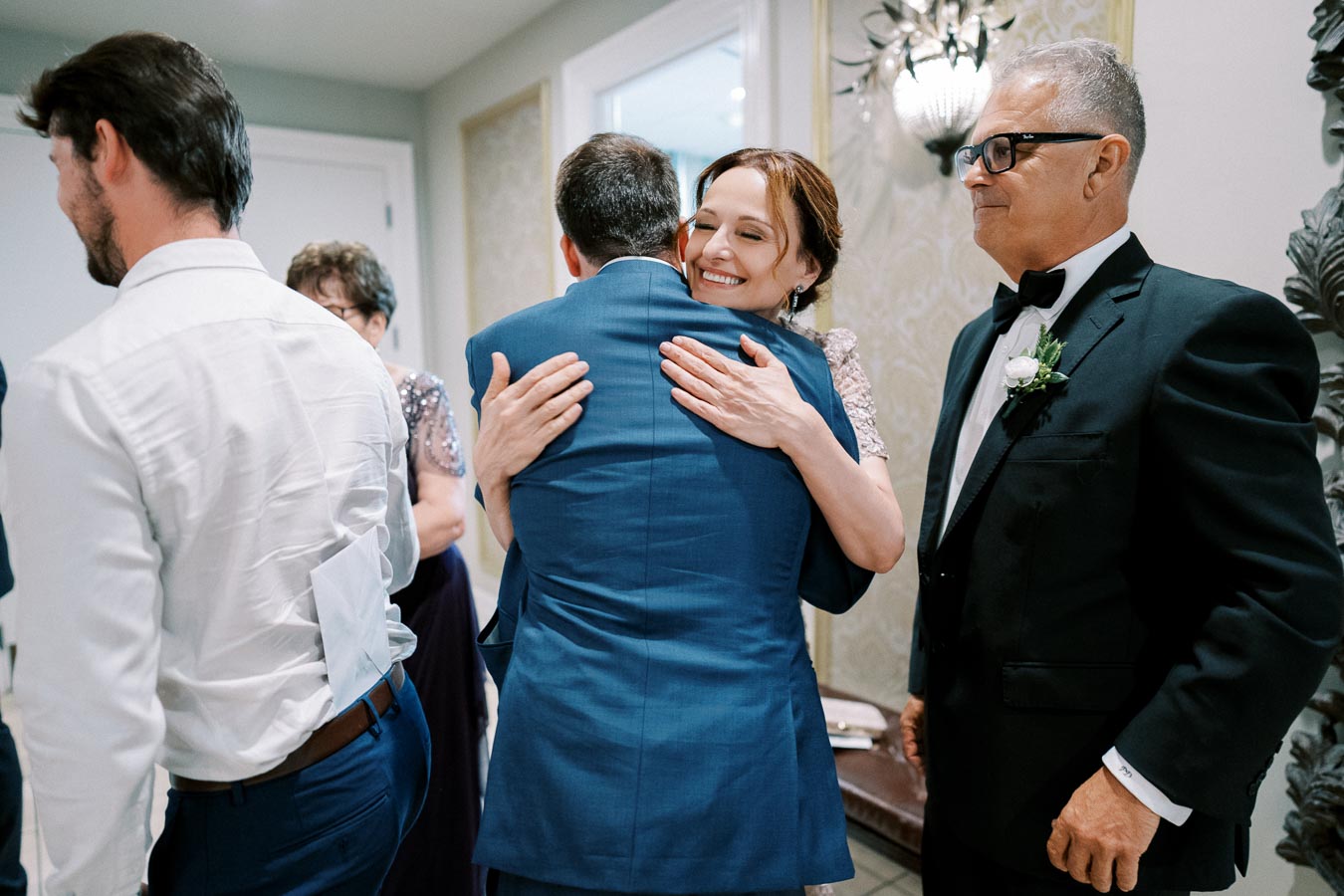 A woman joyfully hugs a man in a blue suit at a formal event, surrounded by other elegantly dressed guests.