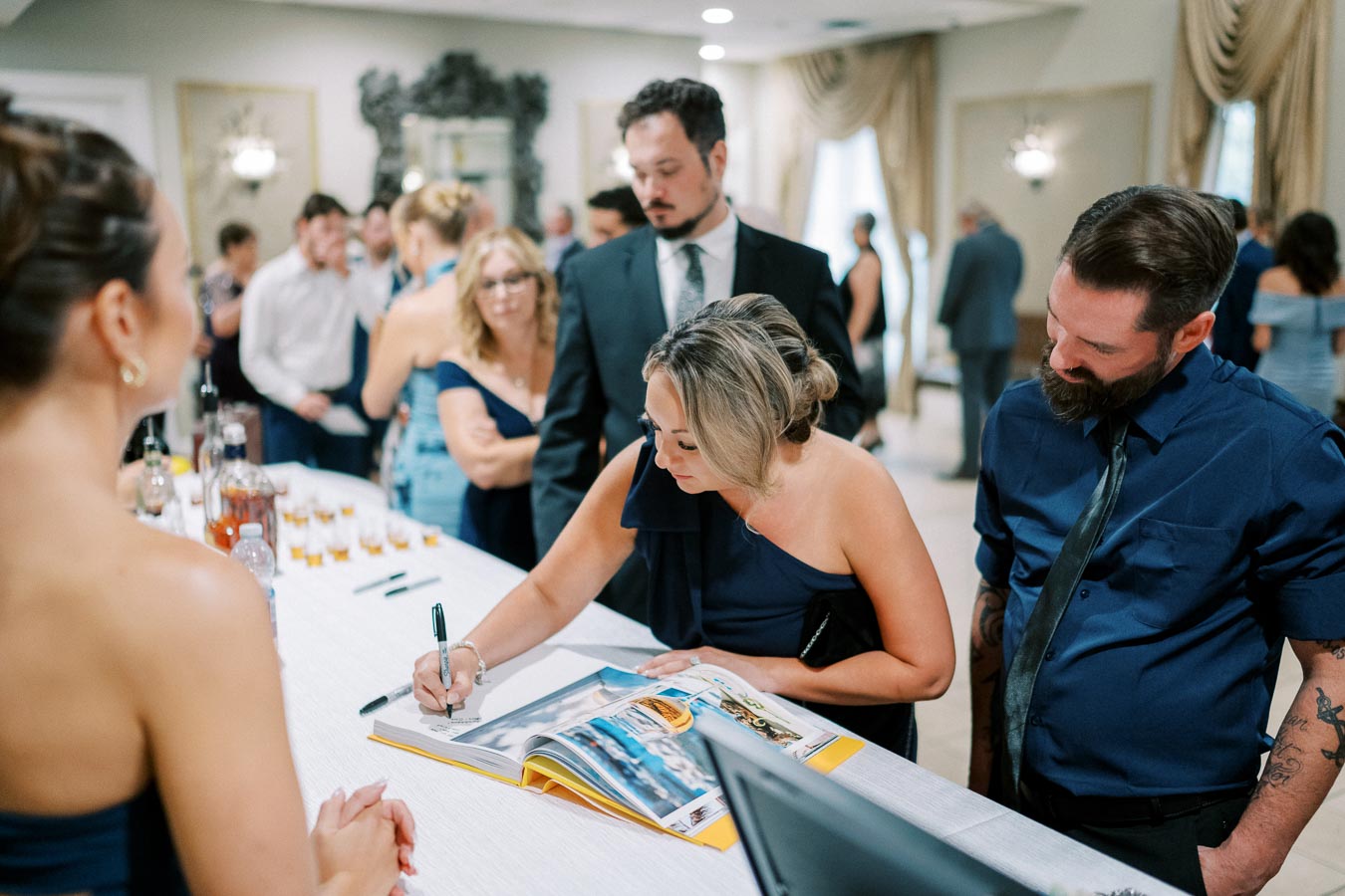 A group of elegantly dressed people in a formal setting, with a woman signing a book at a reception table while others look on.