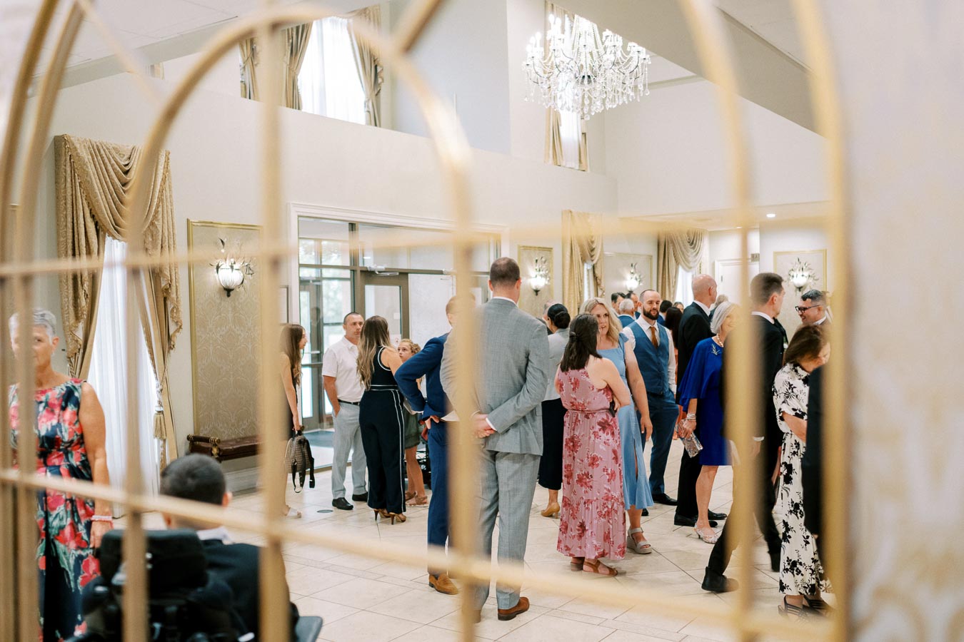 A group of elegantly dressed people socializing and mingling at a sophisticated indoor event with luxurious chandeliers, gold decor, and cream drapery in the background.
