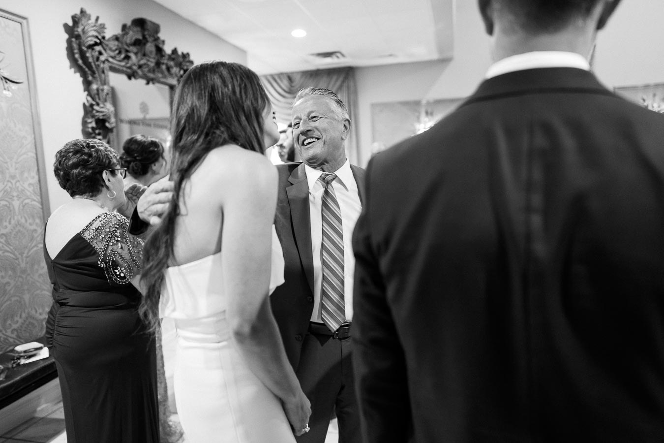 A joyful gathering at a formal event, with a man in a suit smiling warmly at a woman in a strapless dress, surrounded by elegantly dressed guests engaging in conversation.