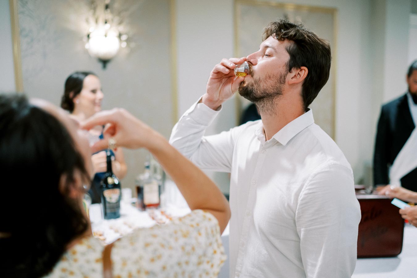 A group of people enjoying drinks at a social gathering, with a man in a white shirt taking a shot in the foreground and bottles on a table in the background.