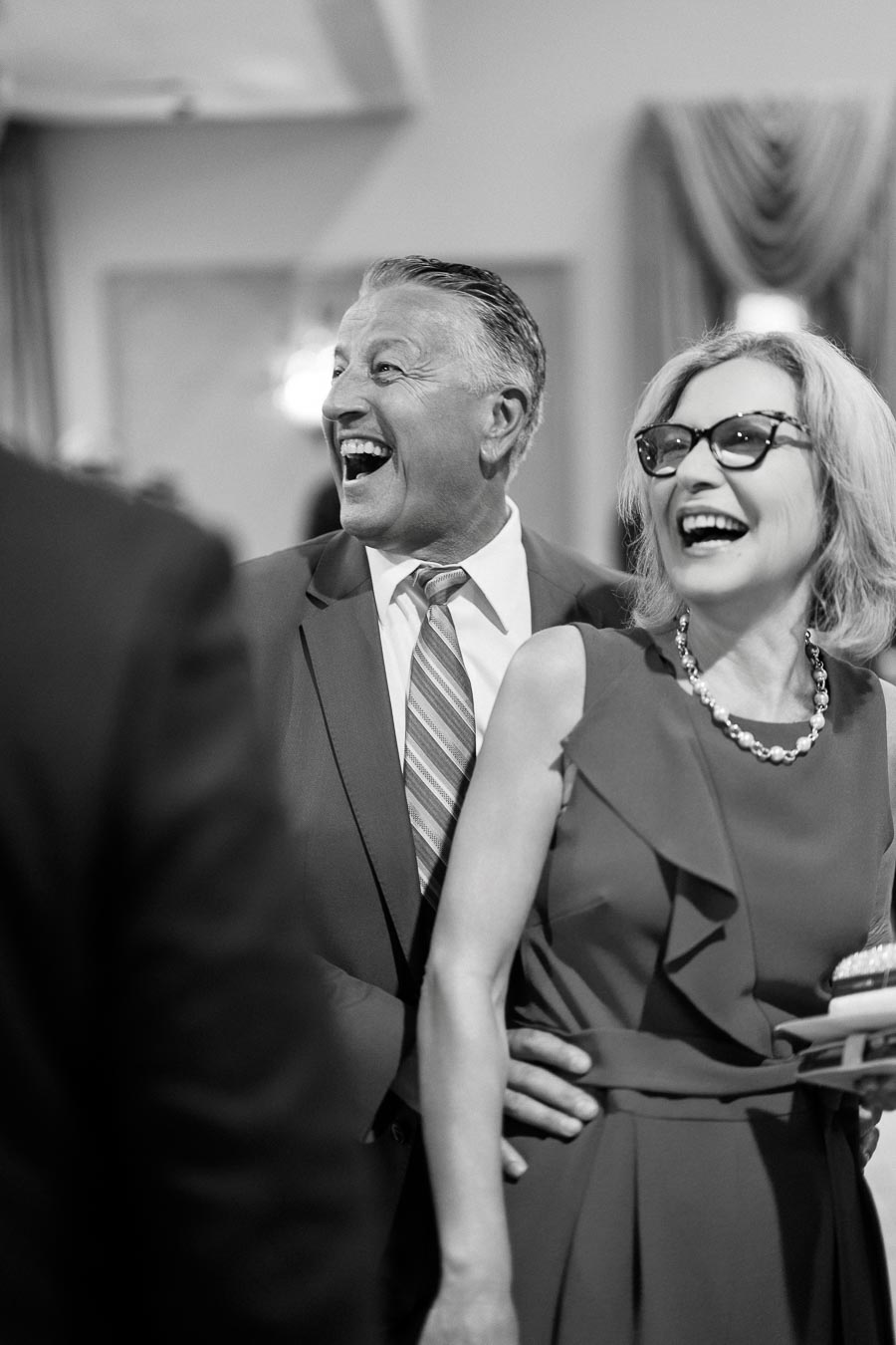 Elderly couple laughing joyfully at an indoor event, dressed in formal attire with man in a suit and woman in a sleeveless dress and glasses. Black and white photograph captures a candid moment of happiness and celebration.