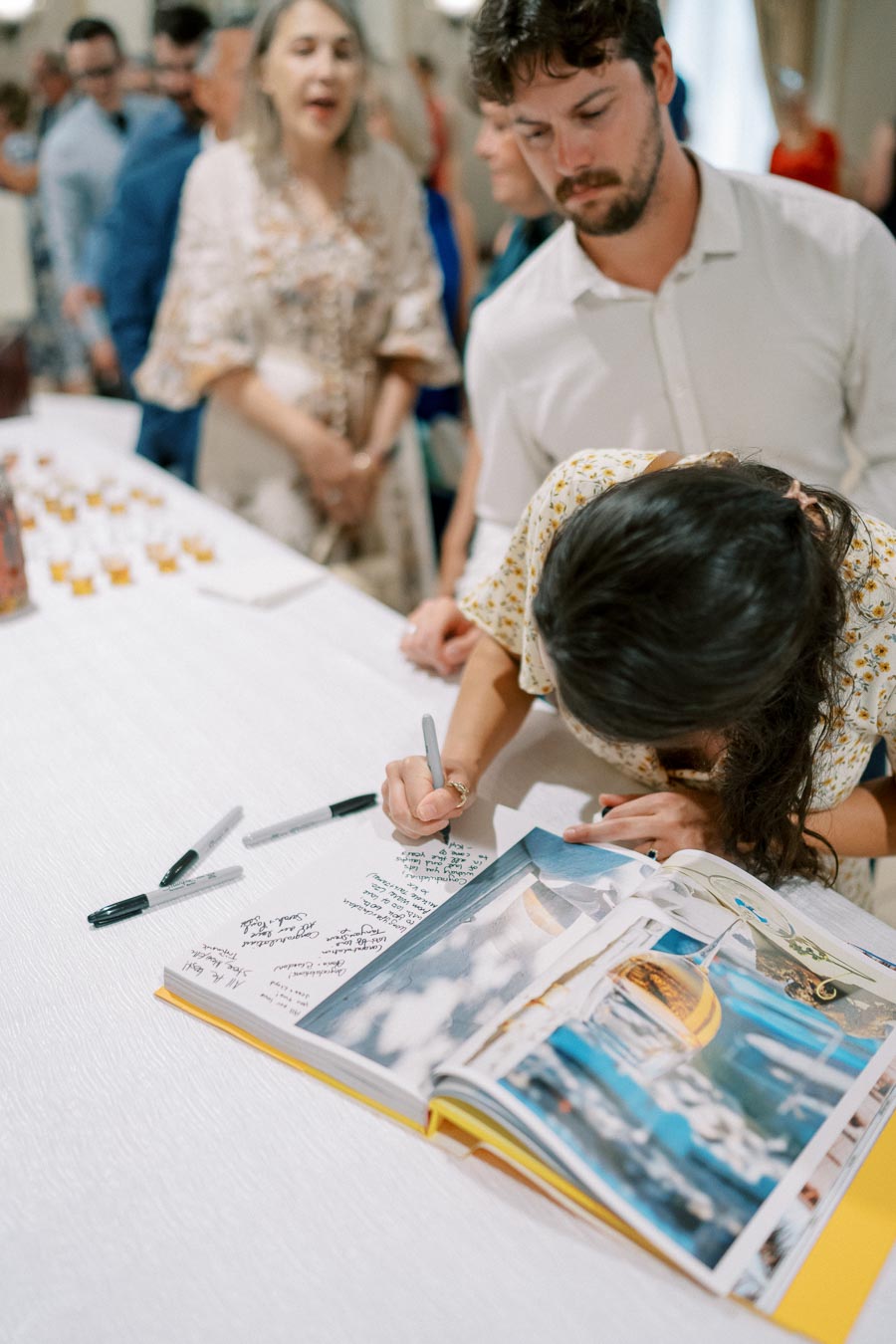 Person signing a guestbook at an event, surrounded by a decorative table with multiple pens and attendees in the background.