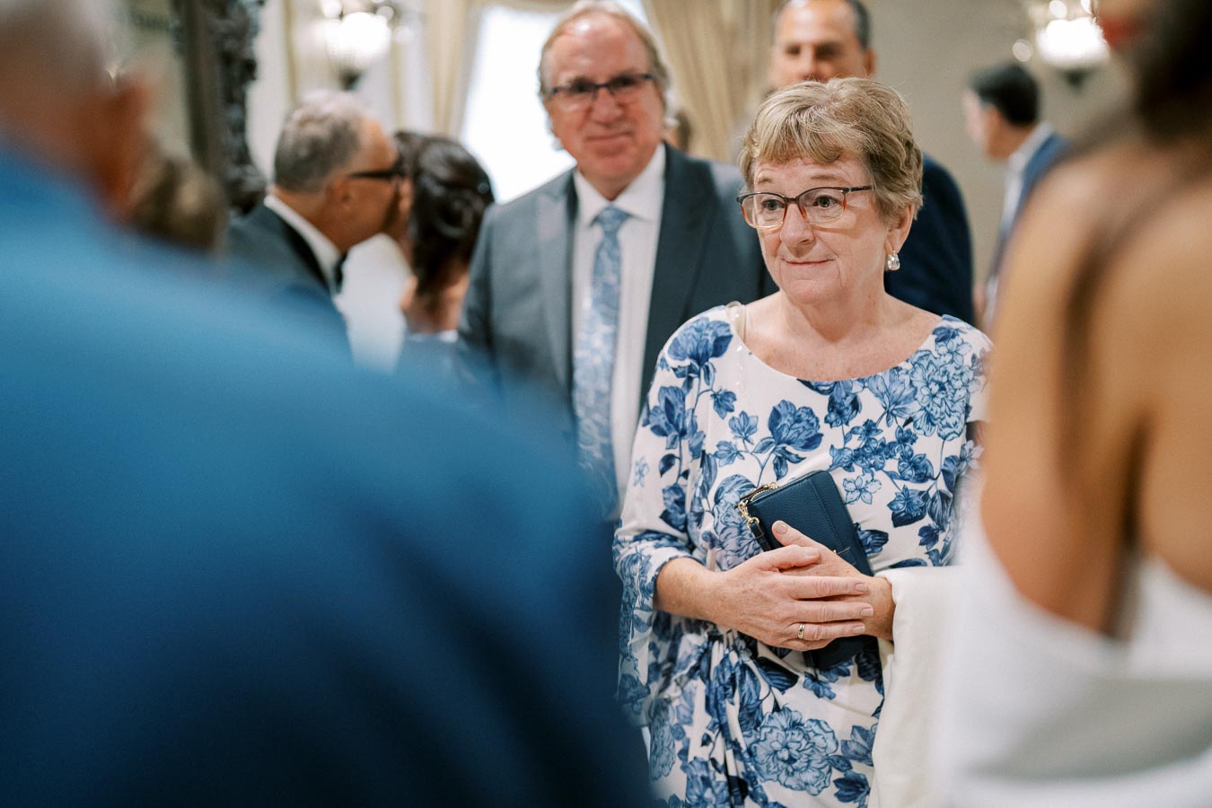 Elderly woman in floral dress holding a clutch at a social gathering, surrounded by other guests in a warmly lit room.