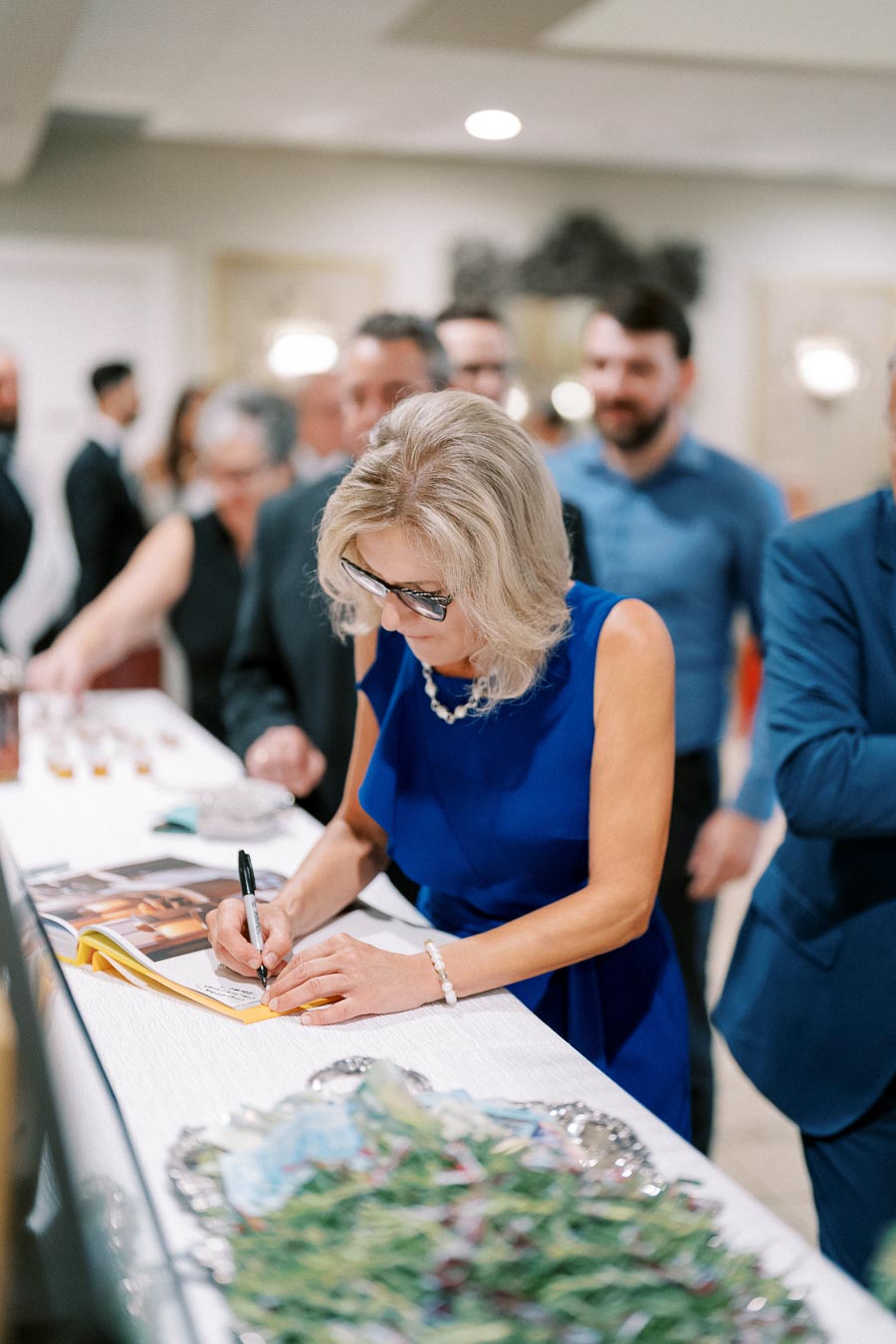 A woman in a blue dress signs a book at a formal event, surrounded by a crowd of elegantly dressed people.