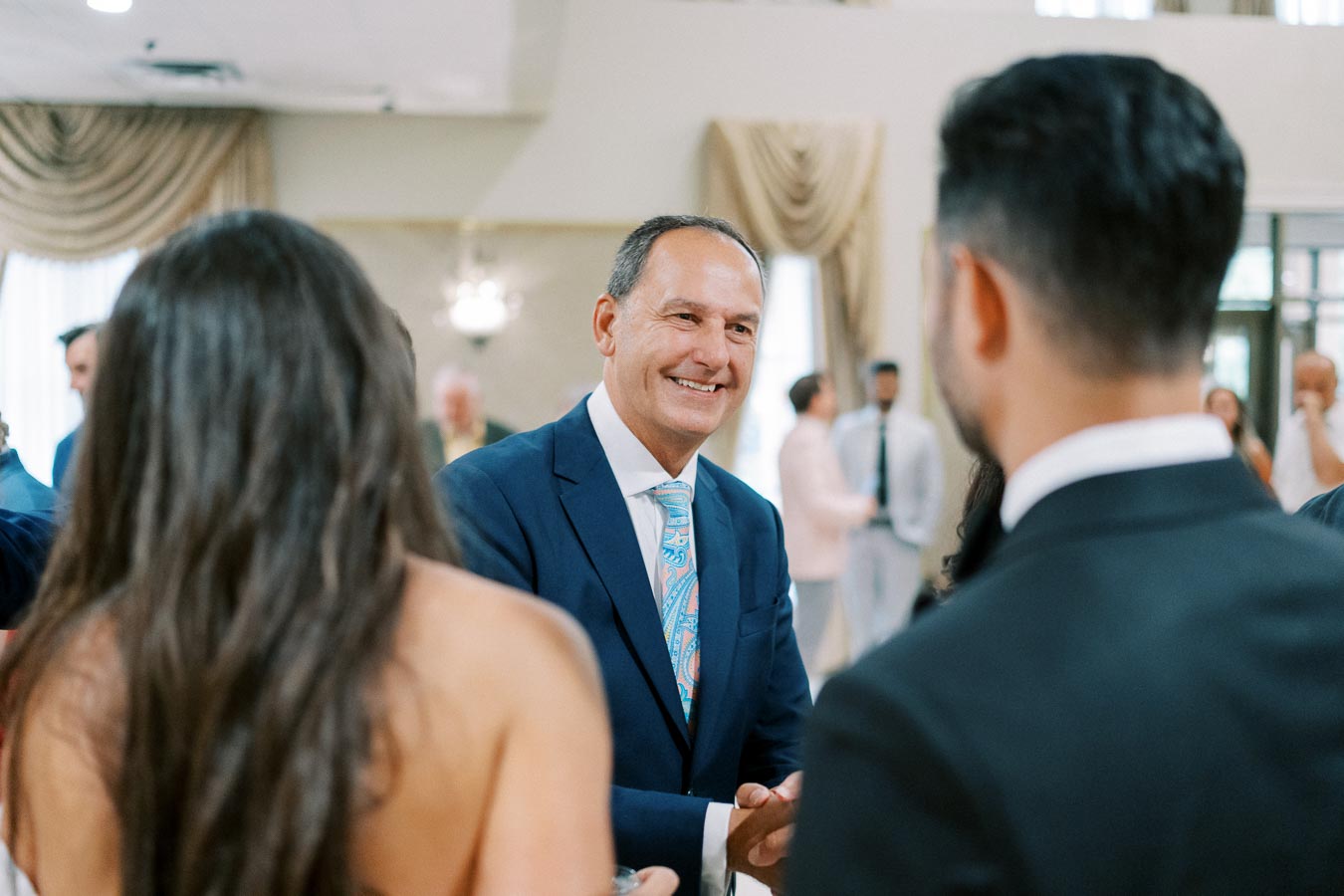 A man in a blue suit smiling and shaking hands at a formal event, surrounded by well-dressed attendees in a banquet hall setting.