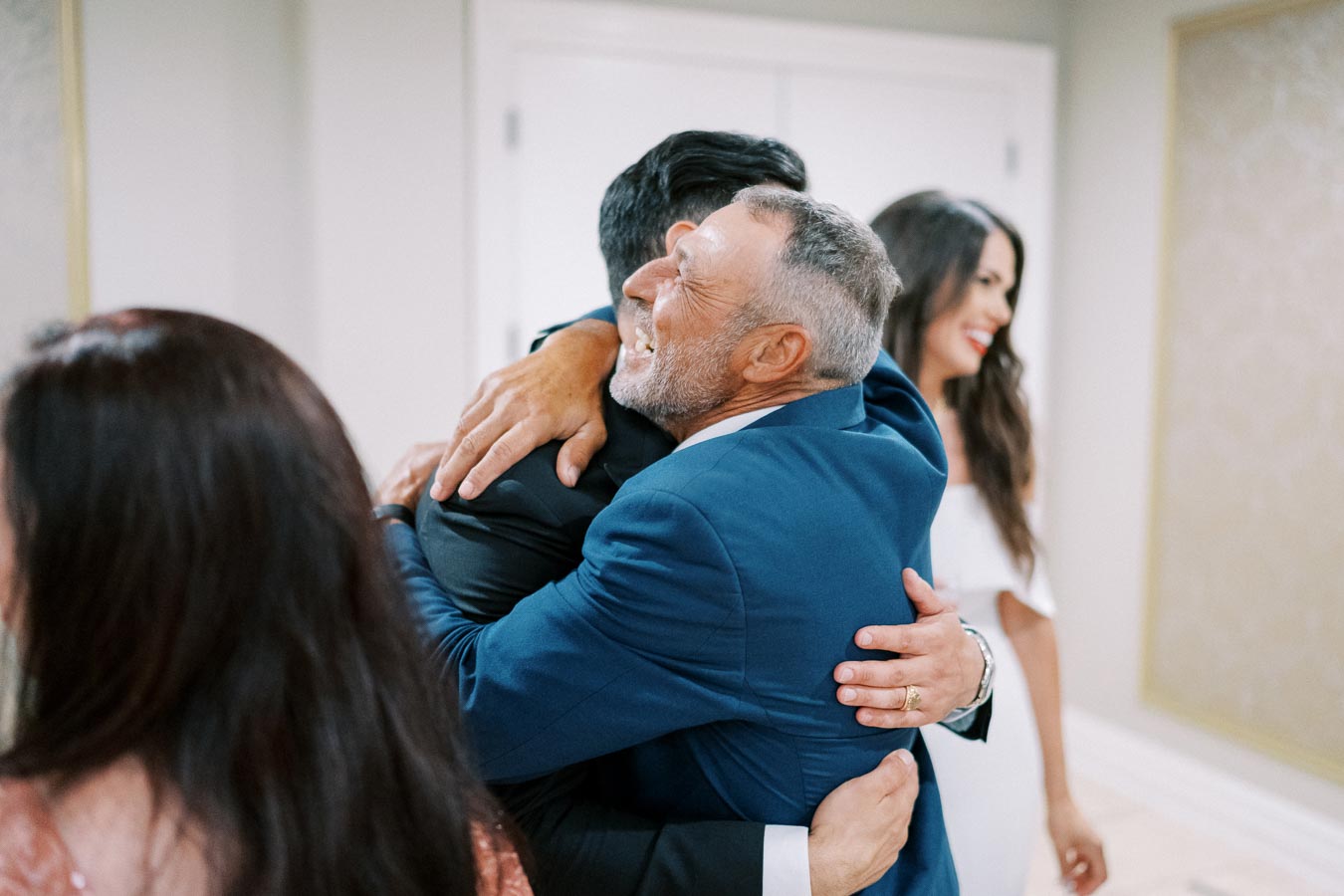 A joyful reunion as two men in suits embrace warmly, capturing a moment of happiness and connection, with a woman smiling in the background.