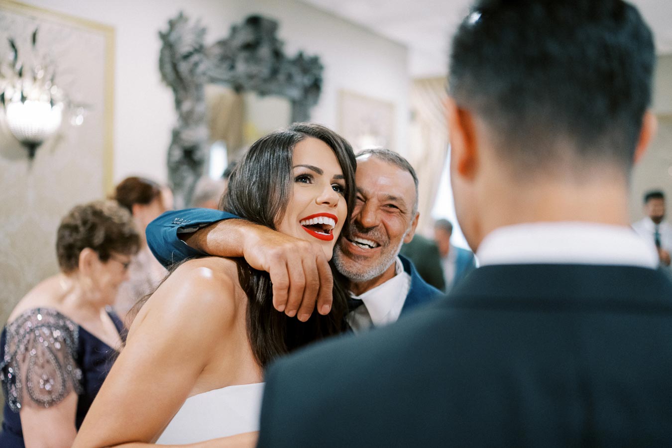 Wedding celebration with a smiling bride embraced by a joyful guest, capturing a candid and happy moment at a reception with elegantly dressed attendees in the background.