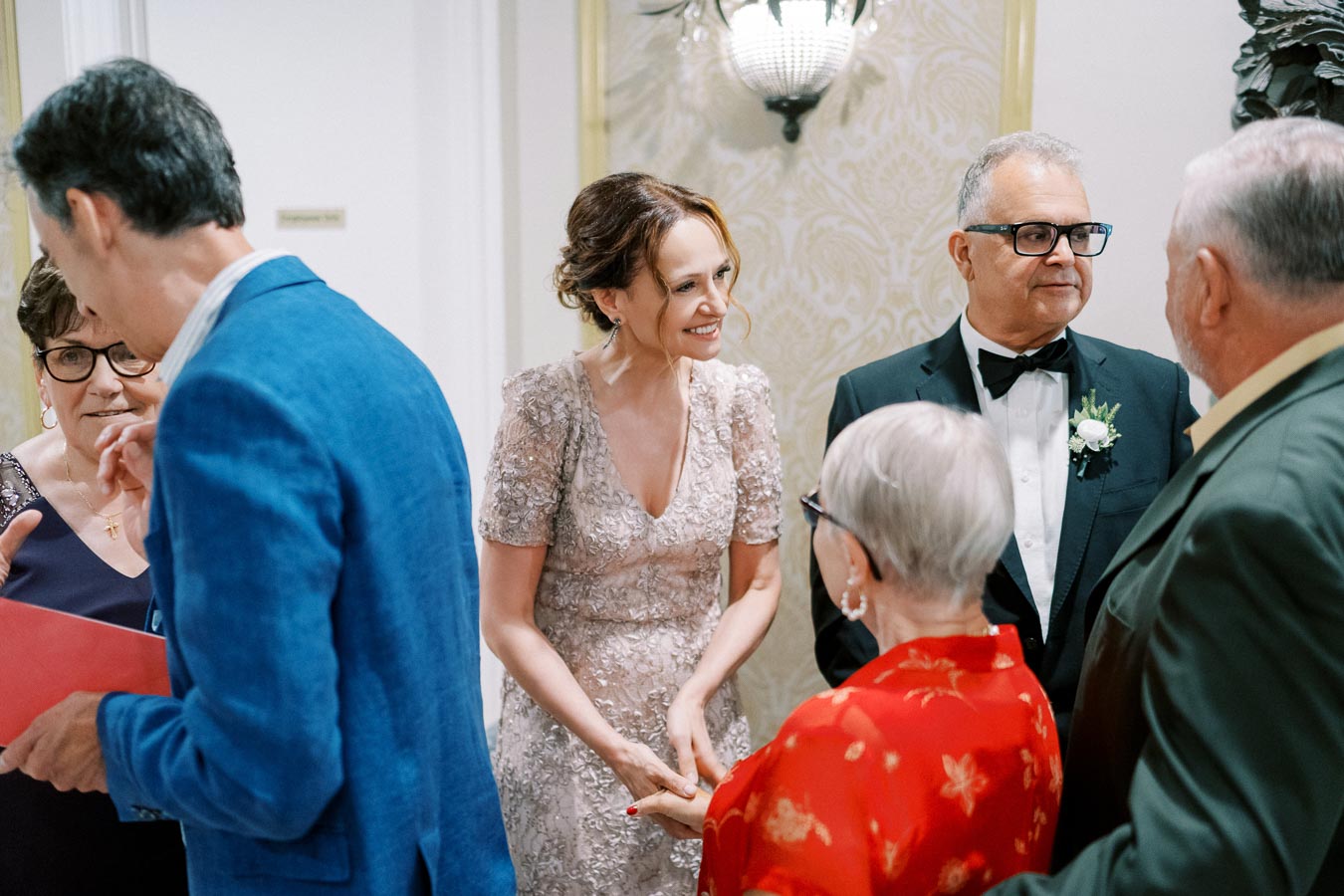 Group of elegantly dressed people socializing at formal event, woman in sparkling dress smiling, man in tuxedo with bow tie nearby.