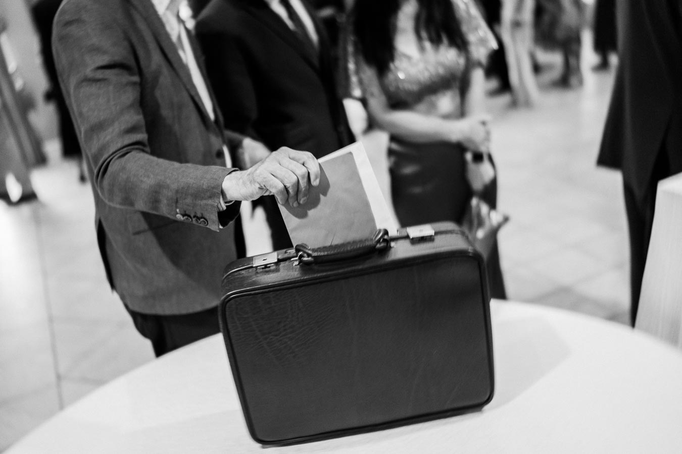 A person in a suit places a document into an open briefcase on a table during a formal event, with other people in the background, captured in black and white.