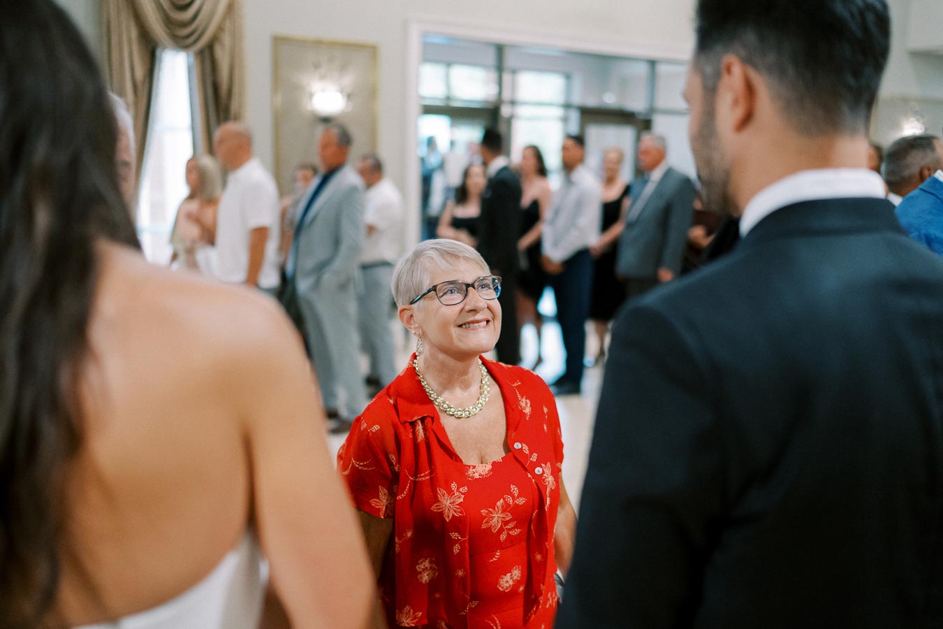 Elderly woman smiling at formal event, wearing a red floral dress, surrounded by people in elegant attire.