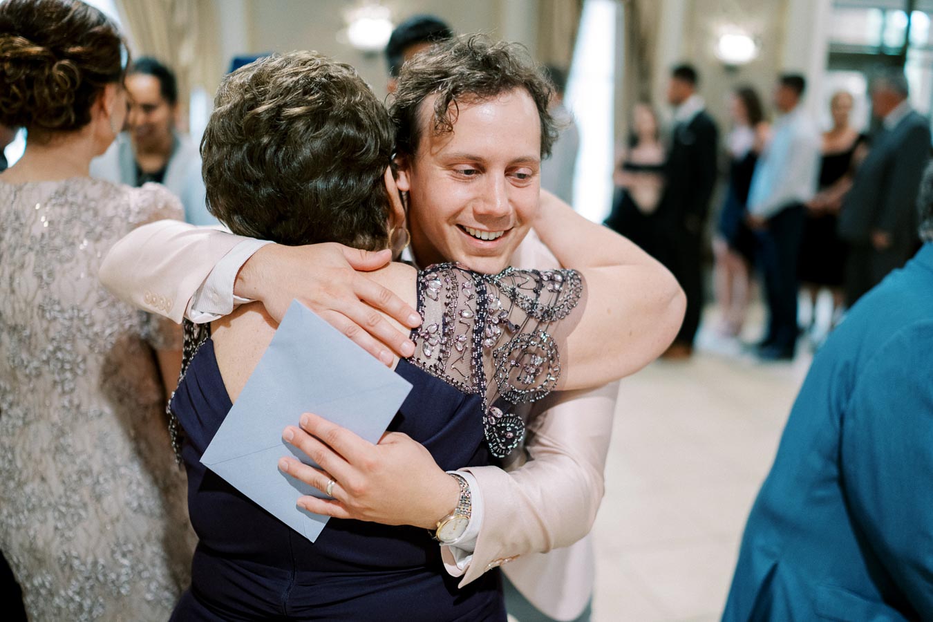 A joyful man embracing a woman at a gathering, with a heartfelt smile on his face, holding a blue envelope; blurred people in formal attire in the background.