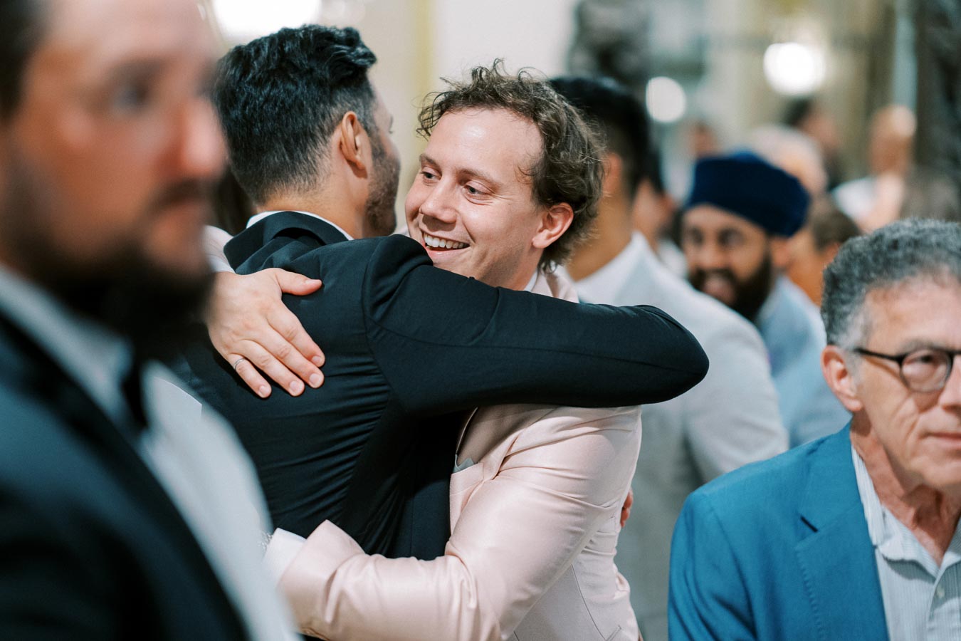 A joyous gathering at a wedding celebration, featuring two men in suits embracing warmly amidst a crowd of guests.