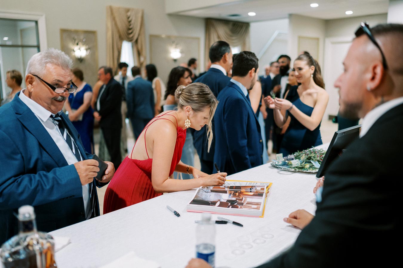 Guests gathered at an elegant event, with a woman in a red dress signing a book at a reception desk, surrounded by people in formal attire.