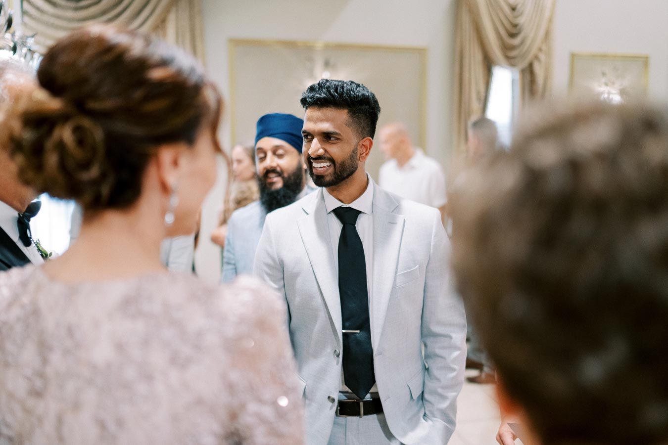 A well-dressed man in a light suit smiles while engaging in conversation at a social gathering. Other guests are visible in the softly focused background, creating a lively, elegant atmosphere.