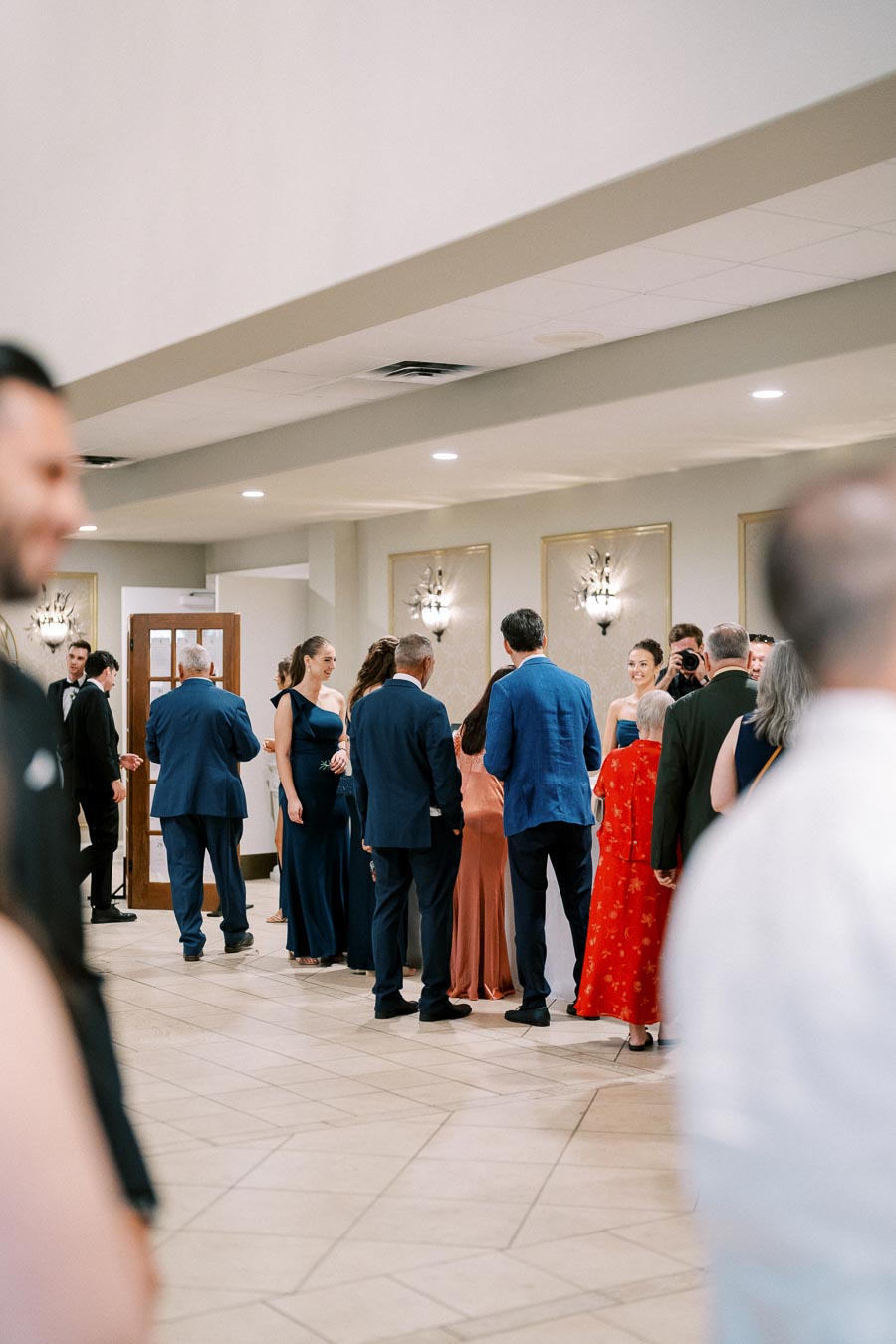 A group of elegantly dressed people in formal attire gathers in a well-lit reception area at an event or wedding.