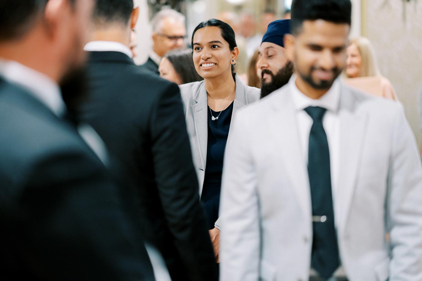 A group of professionally dressed people socializing at a networking event, with a woman in a gray blazer smiling in the center, engaging in conversation.